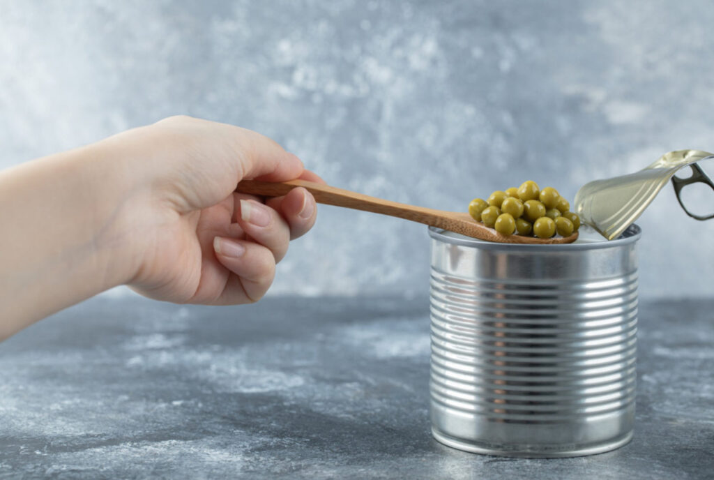 Frau, die grüne Erbsen von der Dose mit Löffel über grauem Tisch nimmt